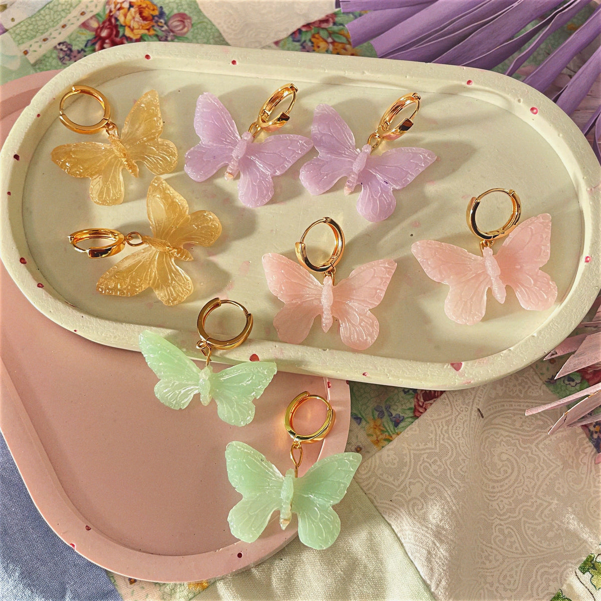 Handmade butterfly-shaped statement earrings crafted with polymer clay, featuring vibrant colors of pink, purple, gold, and green, displayed on trays.