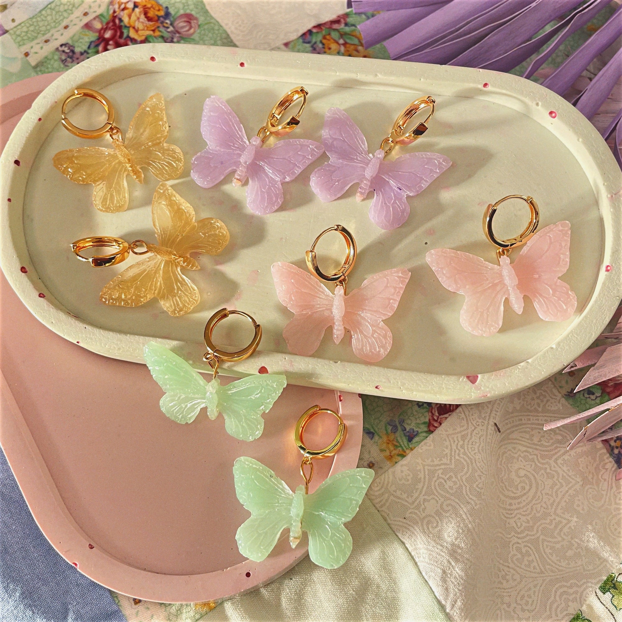 Handmade butterfly-shaped statement earrings crafted with polymer clay, featuring vibrant colors of pink, purple, gold, and green, displayed on trays.